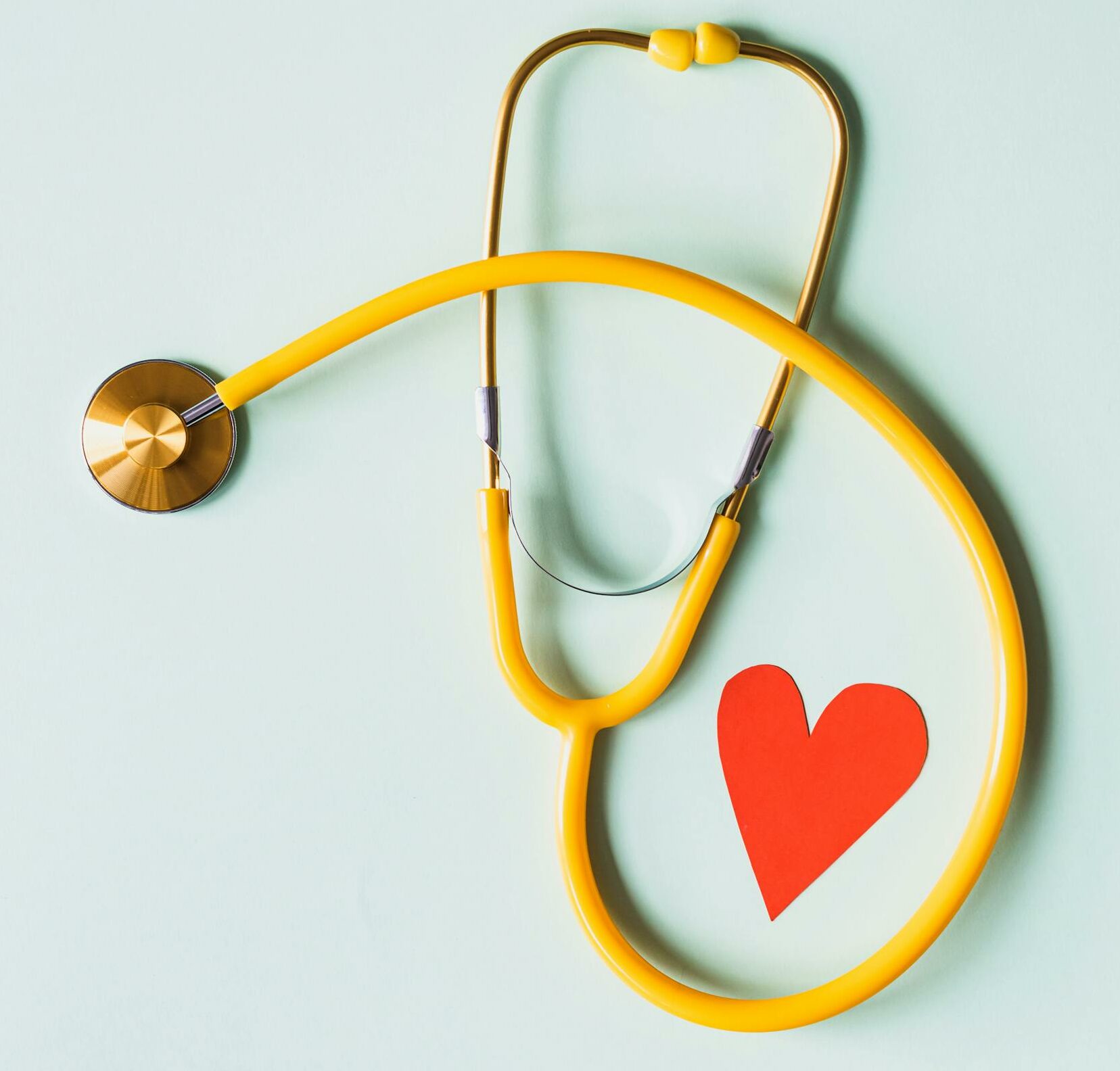 Yellow stethoscope arranged with a red heart cutout on a light green background, symbolizing heart health.